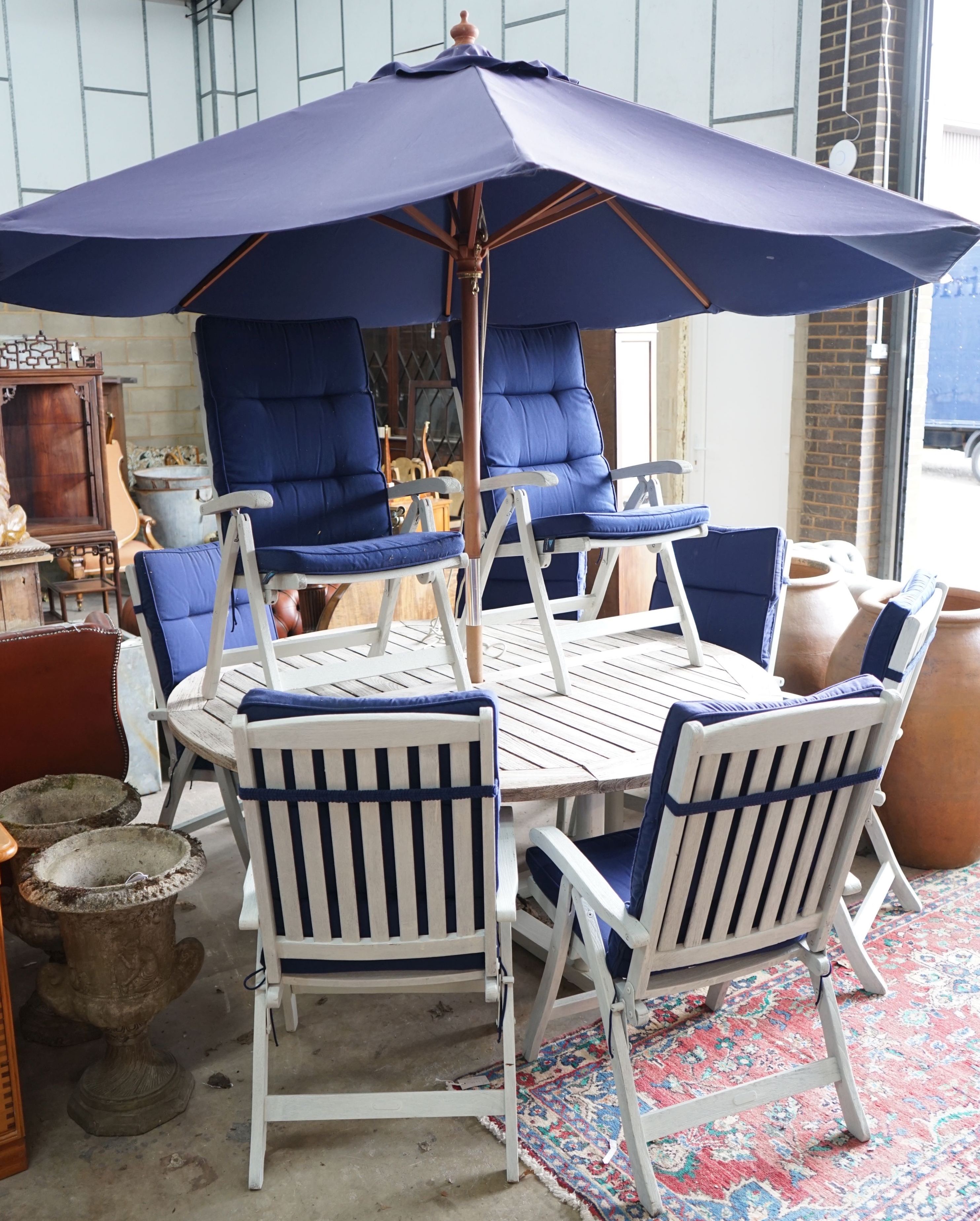 A large circular weathered teak garden table, diameter 180cm, height 72cm, eight painted teak folding chairs with seat pads and a blue canvas crank parasol